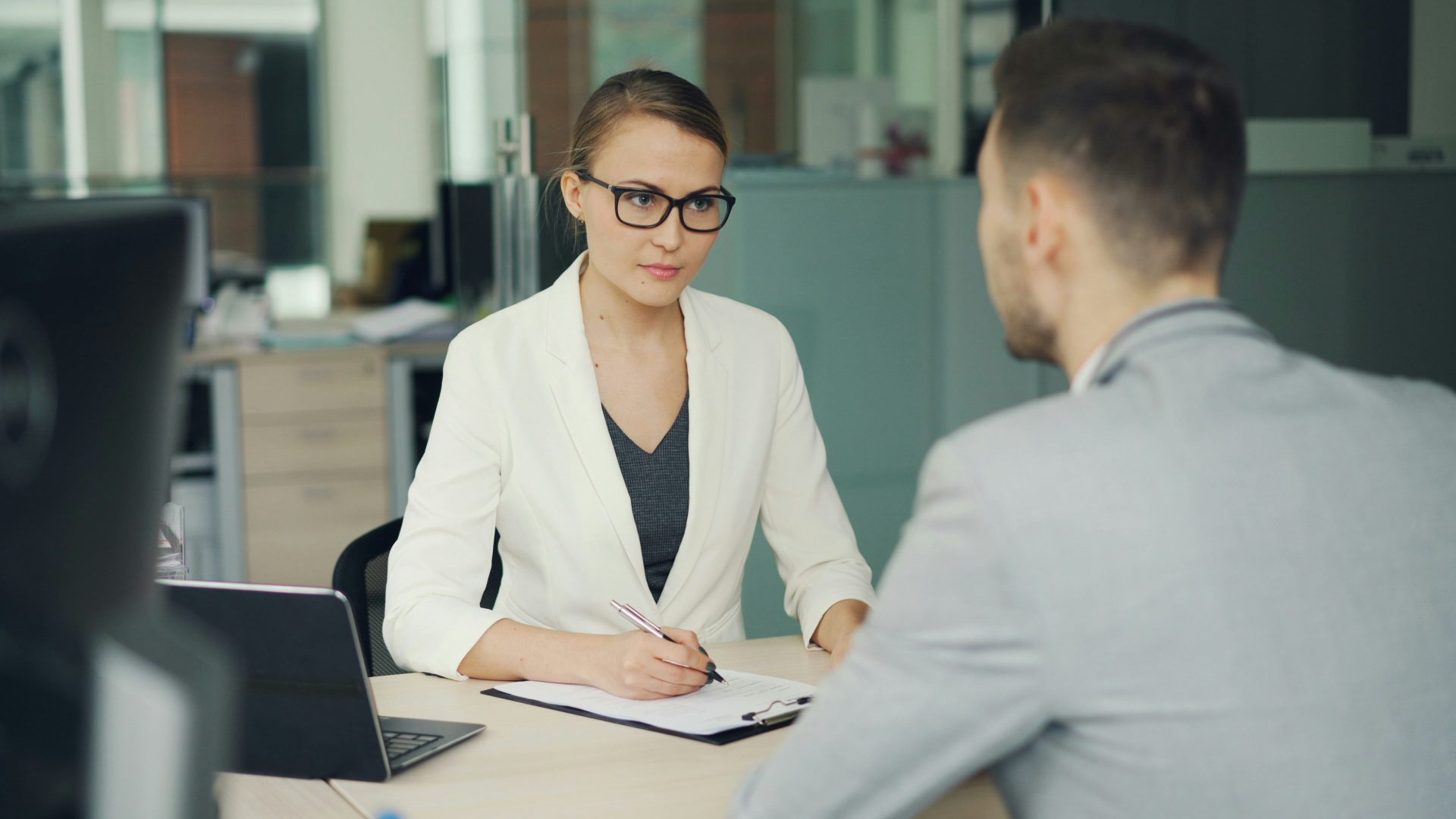 Une femme à lunettes interviewe un homme à son bureau.
